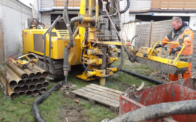 Ein Arbeiter in orangefarbener Arbeitskleidung bedient eine große Bohrmaschine auf einem Grundstück. Neben ihr liegen Rohre, und es gibt eine rote Abfallmulde sowie Schläuche im Umfeld. Im Hintergrund sind Wände und ein Zaun sichtbar.