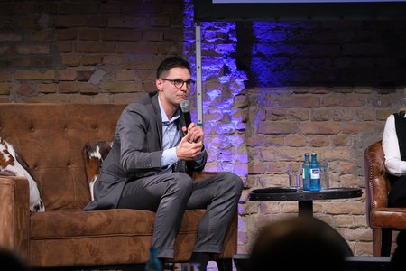 Ein Mann in einem grauen Anzug sitzt auf einer Couch und hält ein Mikrofon in der Hand. Er wirkt nachdenklich und spricht zu einem Publikum. Im Hintergrund sind Steine und eine weitere Person auf einem Stuhl zu sehen.