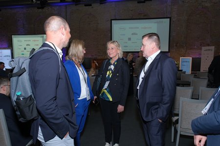 Vier Personen in formeller Kleidung stehen in einem modernen Veranstaltungsraum im Gespräch. Eine Frau trägt eine bunte Bluse, während die anderen Männer Anzüge tragen. Im Hintergrund sind Veranstaltungsbanner und Stuhlreihen sichtbar.