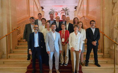 Gruppensfoto von 15 Personen auf einer Treppe in einem eleganten Gebäude. Die Erwachsenen stehen in zwei Reihen, lächelnd und formell gekleidet. Im Hintergrund sind große Fenster und Palmen sichtbar.