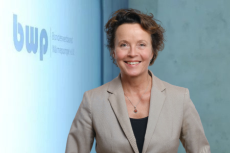 Eine Frau mit lockigem Haar trägt einen hellen Blazer und lächelt. Sie steht vor einer Wand mit dem Schriftzug "bwp" und scheint in einem professionellen Umfeld zu sein.